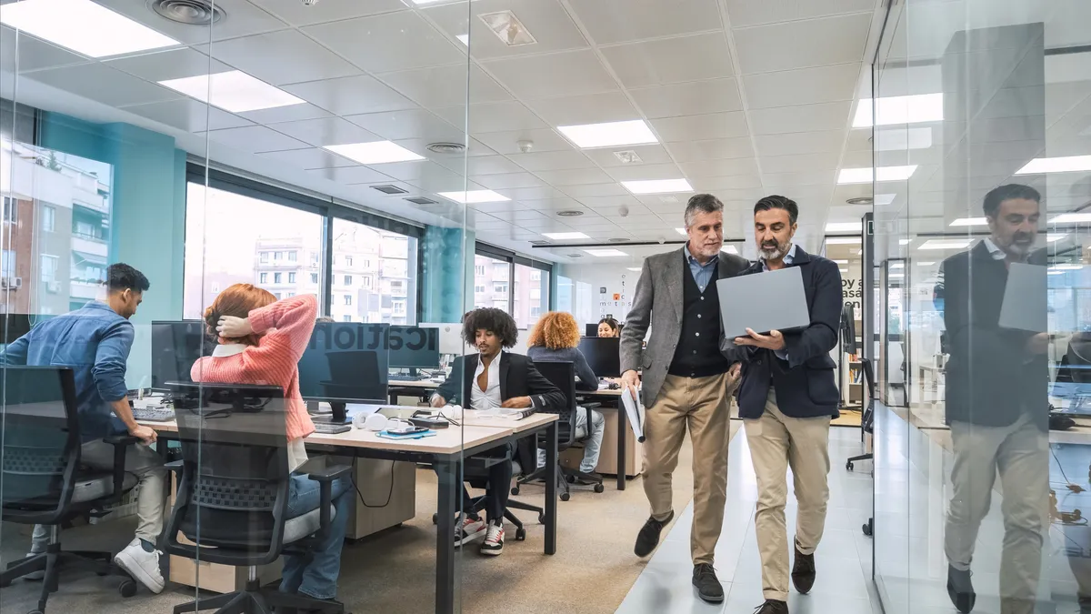 Managers hold a laptop while walking alongside employee desks.