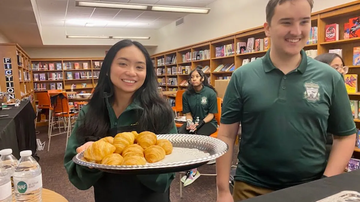 Two high school students stand facing the camera behind a high table. One student is holding a tray of croissants.