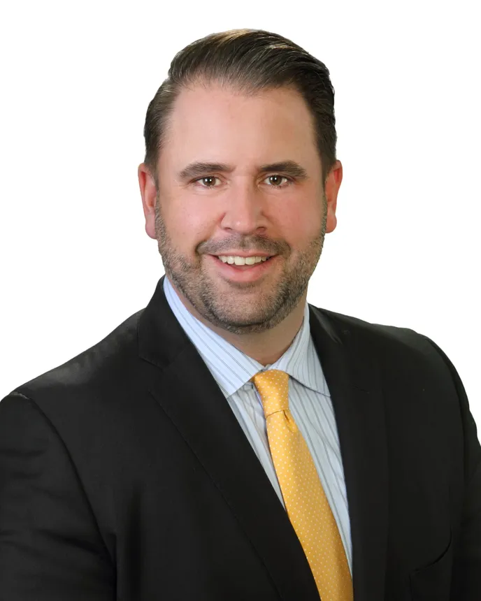 Head and shoulders shot of a man in a dark suit and yellow tie, with a short beard and brown hair.