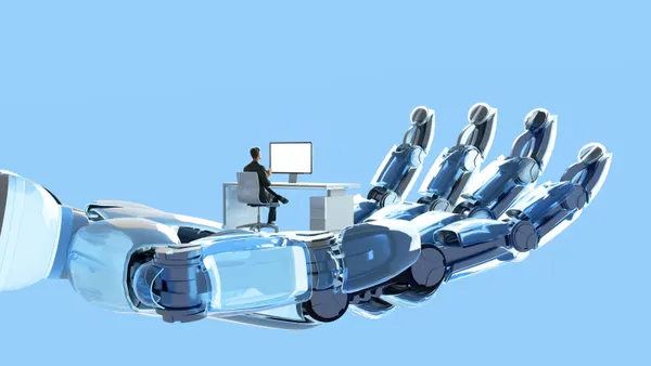 A man sits at a computer desk which is in a giant robot hand.