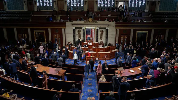 A wide-angle of the floor of the House is seen with people walking around and sitting at desks.
