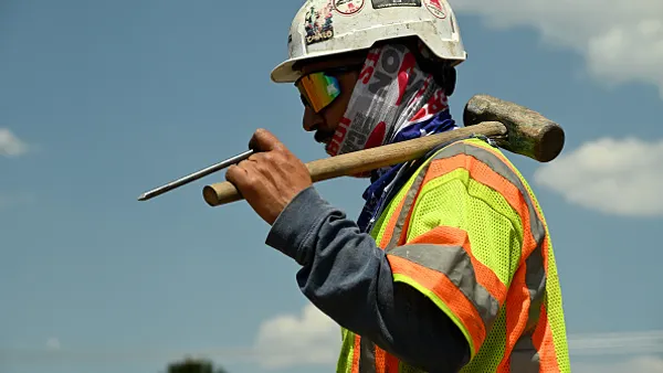 A construction worker with a vest on and a sledgehammer on his shoulder.