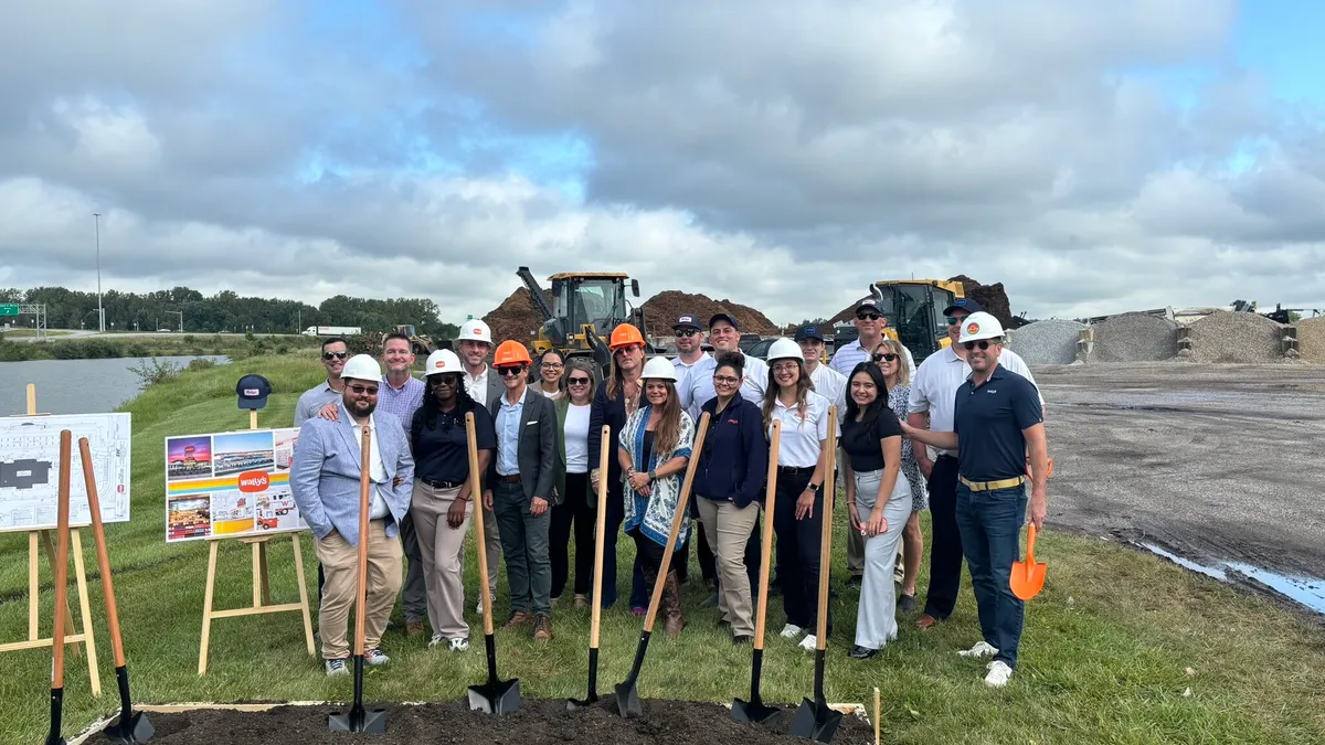 A photo of a number of people standing begind a patch of bare earth with shovels stuck in it in an open field. There is construction equipment behind them. A sign on an easel next to them shows several small pictures of a retail locations. Text in the center fo the sign says "Wally's."