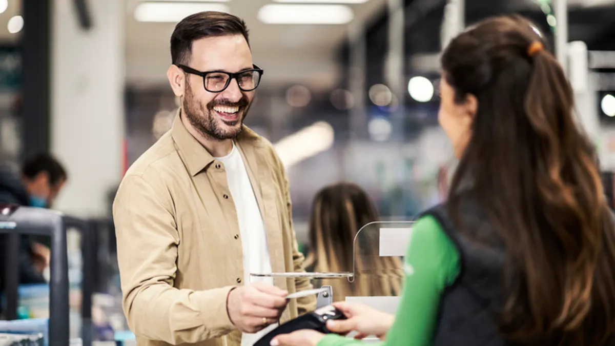 A man with glasses smiling as he pays a cashier in a busy store. The cashier in a green uniform scans his card, conveying a friendly interaction.