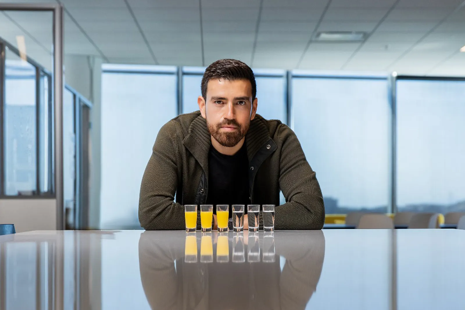 Arturo Elizondo leans on a reflective table with six shot-glasses of liquid in front of him. The three on the left are orange, the three on the right are clear.