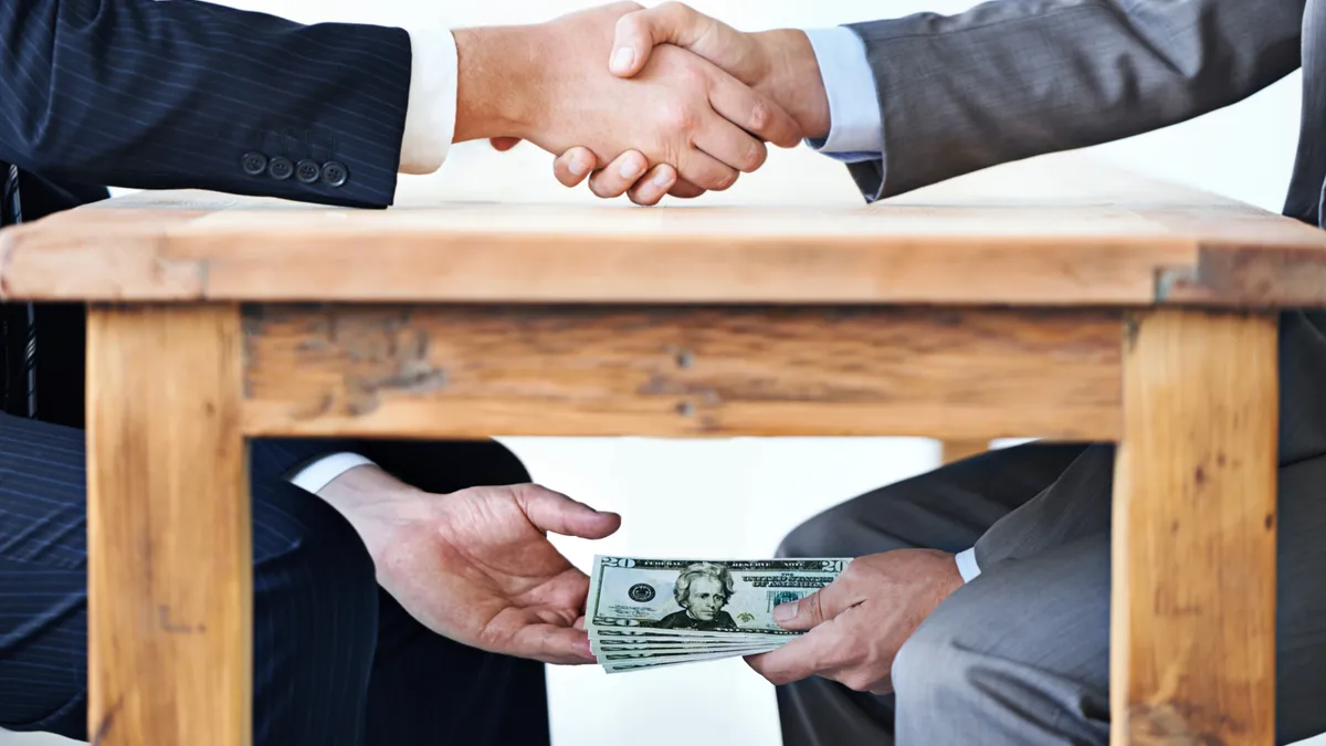 Two people shake hands at a table. Underneath, one passes money to the other.