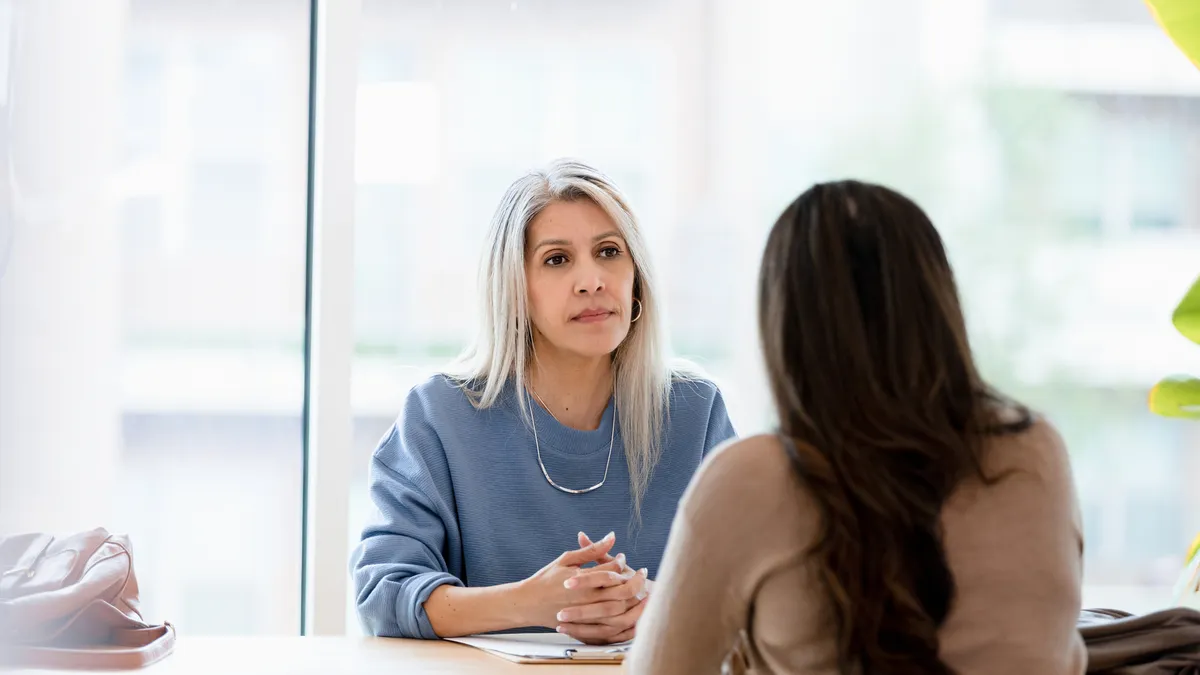 A woman with long gray hair listens to another person speaking.