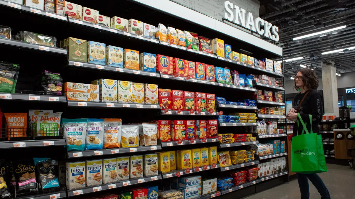 A person with a shopping bag stands in front of a grocery store display labeled snacks.