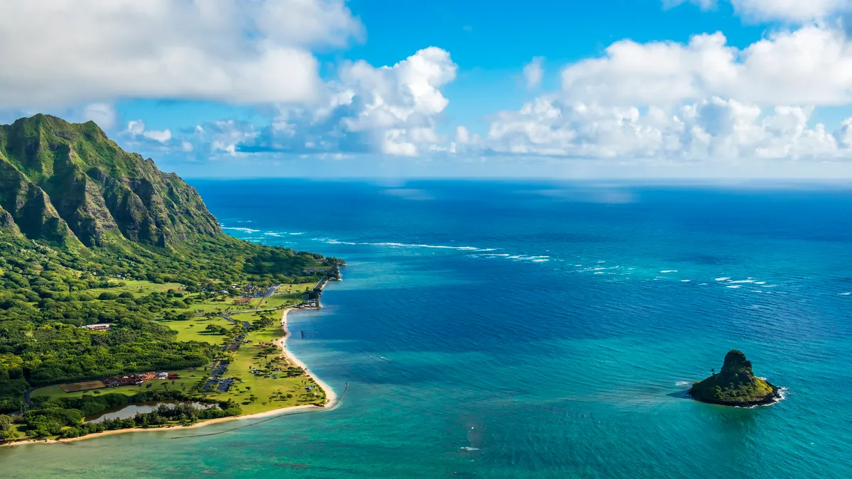 An aerial view of the ocean off of a green island with clouds in the distance.