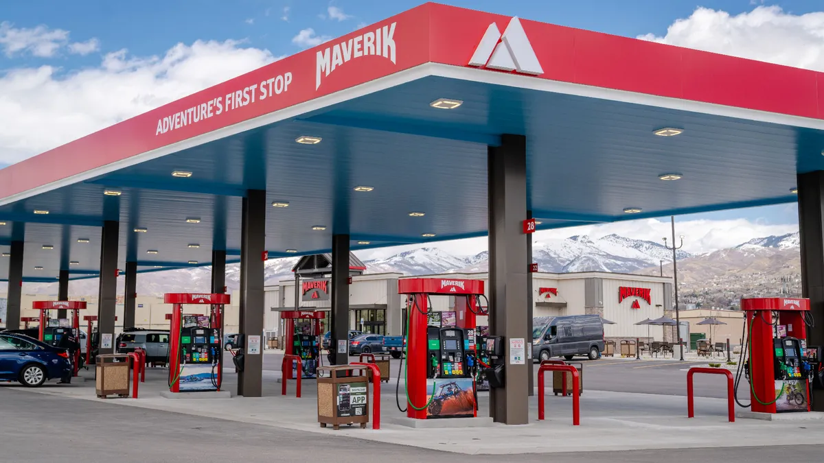 A photo of the exterior of a gas station and convenience store. The fuel canopy is red and reads "Adventure's first stop" and "Maverik" while signs on the gas pumps and stores say "Maverik."