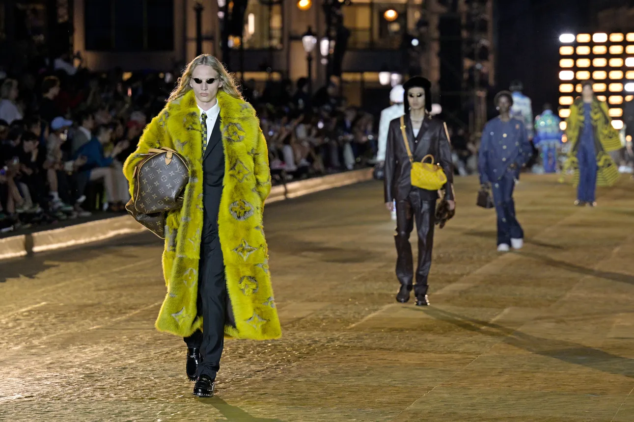 A man in a yellow fur coat walks down a fashion runway wearing sunglasses.