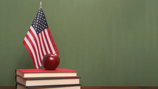 A small American flag is hanging near a pile of books with an apple on top. In the background is a chalkboard
