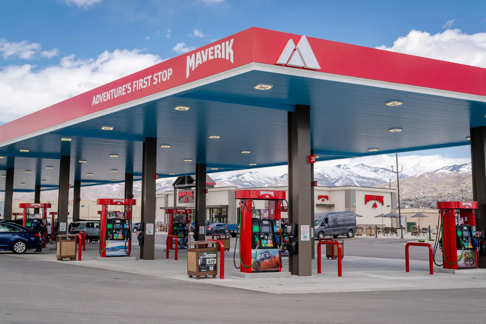 A photo of the exterior of a gas station and convenience store. The fuel canopy is red and reads "Adventure's first stop" and "Maverik" while signs on the gas pumps and stores say "Maverik."