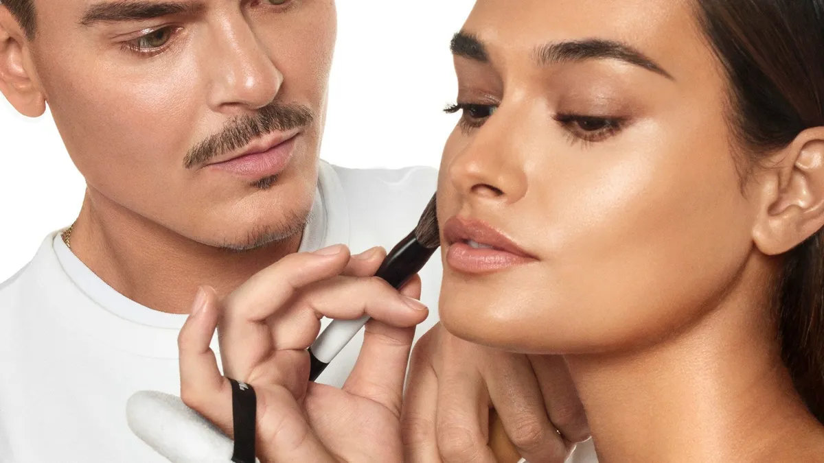 Person with short brown hair holds makeup brush to face of person with long brown hair