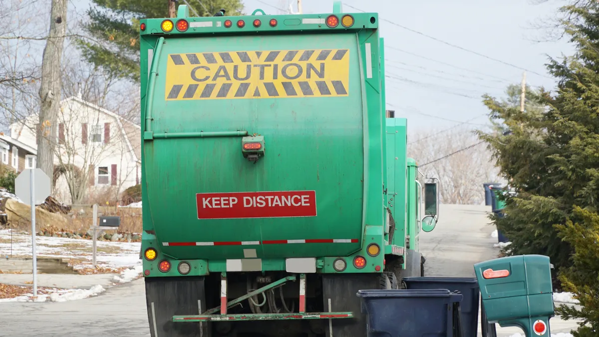 Rear view of a waste collection vehicle, with the word "caution," next to empty collection carts.