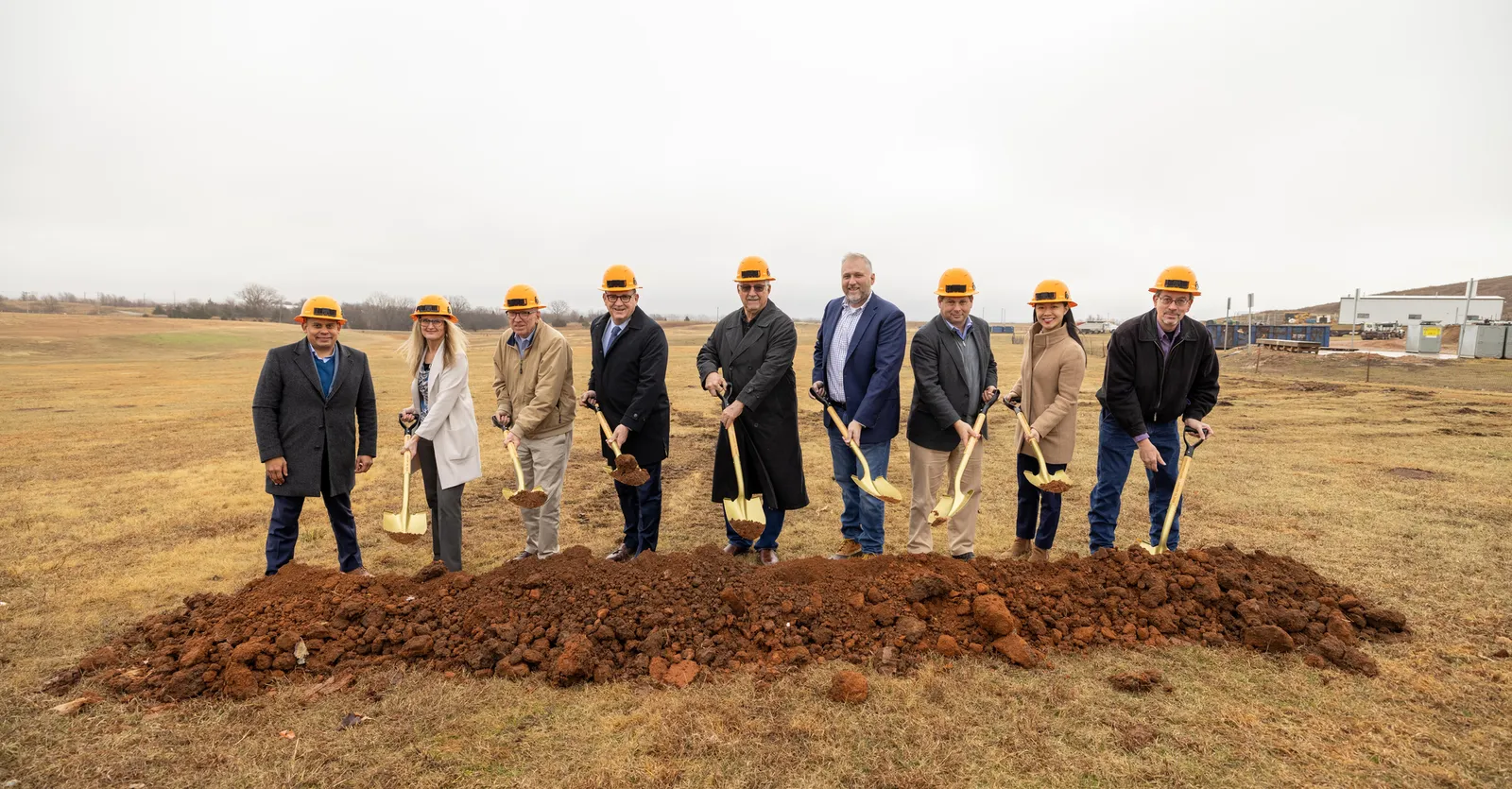 Nine people stand in front of a pile of dirt on a lot covered in yellowed grass in winter.