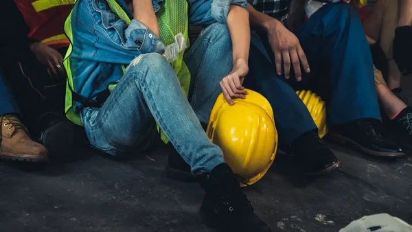 A closeup of several people's legs as they sit on the ground and hold hard hats in their hands.