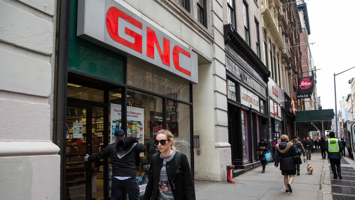 People walking down a city sidewalk past a row of shops.