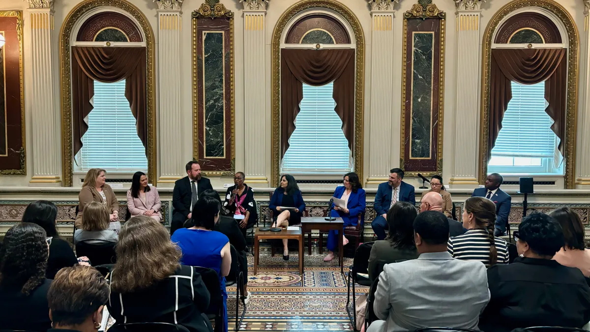 A row of presenters sits in front of rows of attendees in an ornate room.