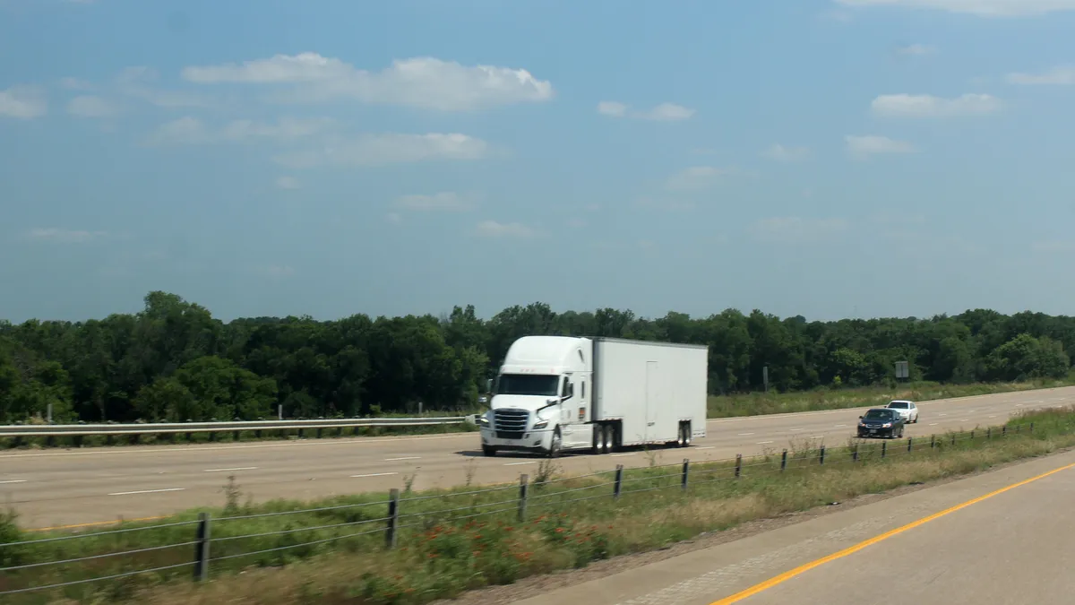 A white tractor-trailer travels in the middle lane of a Texas highway ahead of two cars with green trees, blue skies and white clouds in the background.