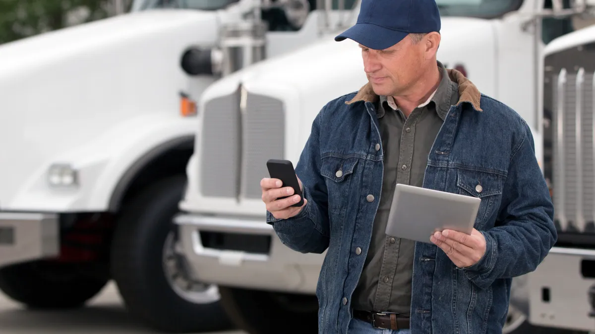 A royalty free image from the trucking industry of a truck driver using a cellphone and tablet computer in front of semi trucks