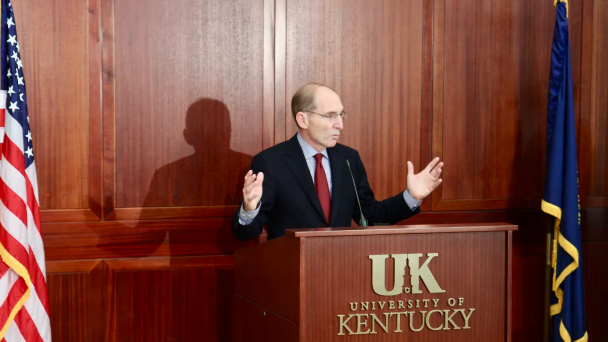 A bald man speaks behind a podium between the U.S. flag and the Kentucky state flag.