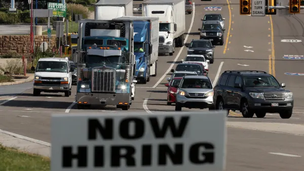 Trucks drive on a road past a "NOW HIRING" sign.