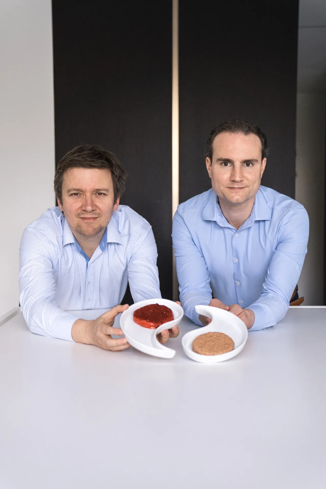 Hermes Sanctorum and Andy de Jong hold plates shaped like a white yin-yang with a patty of raw plant-based meat in each.