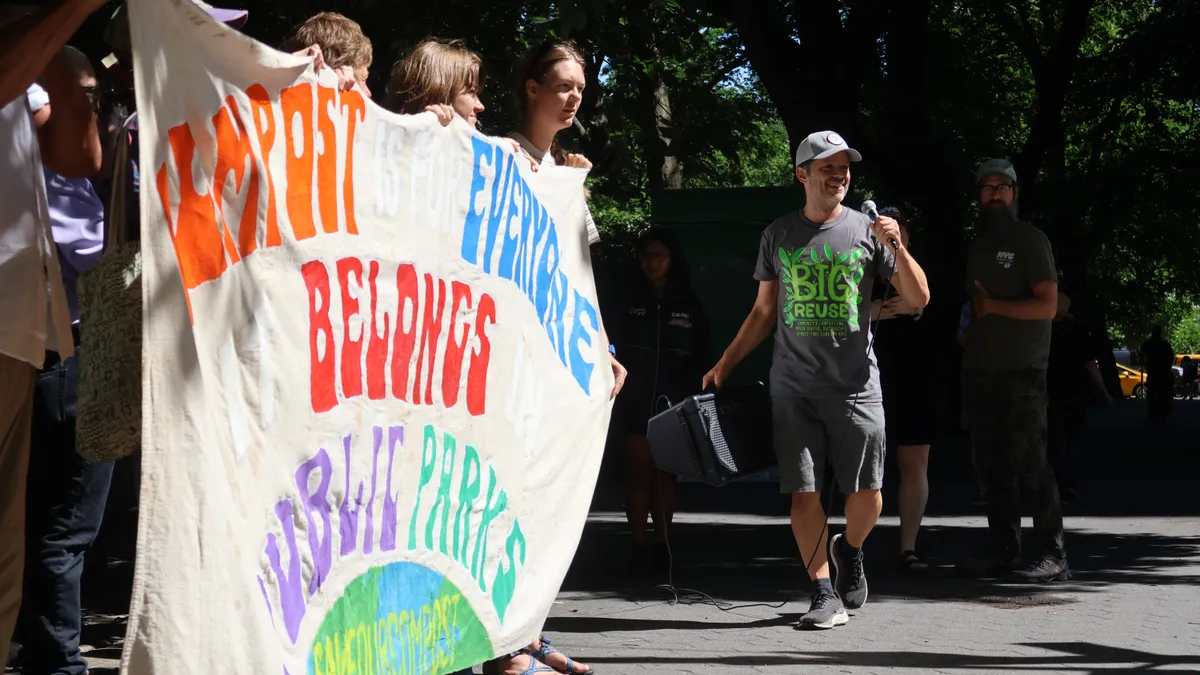 A man walks near a banner holding a microphone and speaker. The banner says "Compost Is For Everyone, It Belongs In Public Parks."