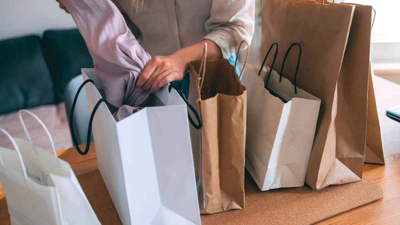 A person pulls items out of one paper shopping bag with handles that is on a table next to four similar bags.