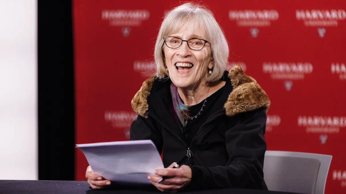 Economist Claudia Goldin speaks enthusiastically at a podium in front of a Harvard-themed backdrop