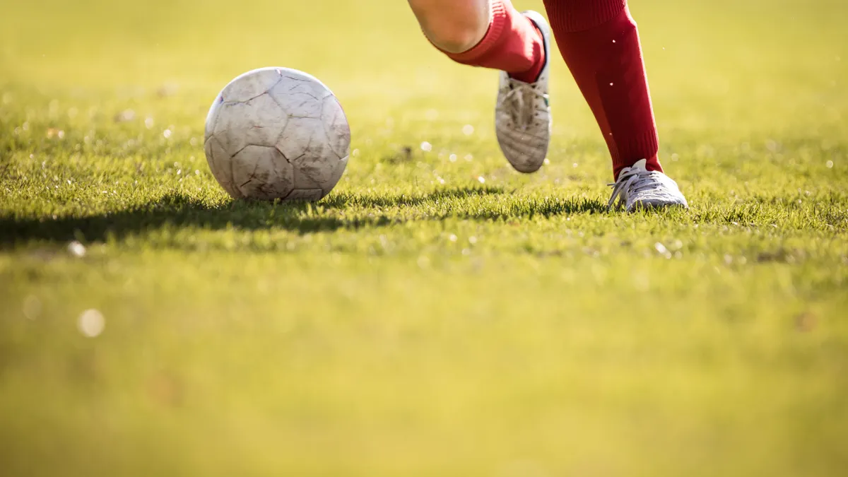 A pair of legs in soccer cleats and red shin guards kicks a soccer ball on a grassy field