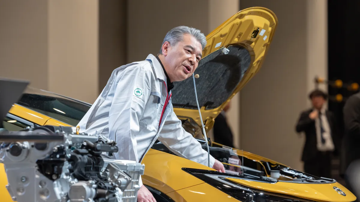 Toyota's chief technology officer, Hiroki Nakajima, speaks at a presentation with a yellow Toyota Prius and engine on display.