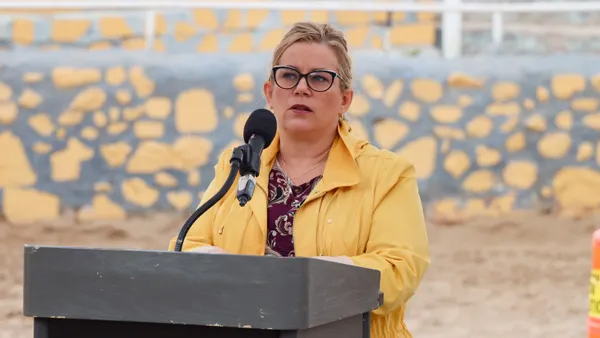 A woman with glasses wearing a yellow jacket speaks into a microphone at a podium.