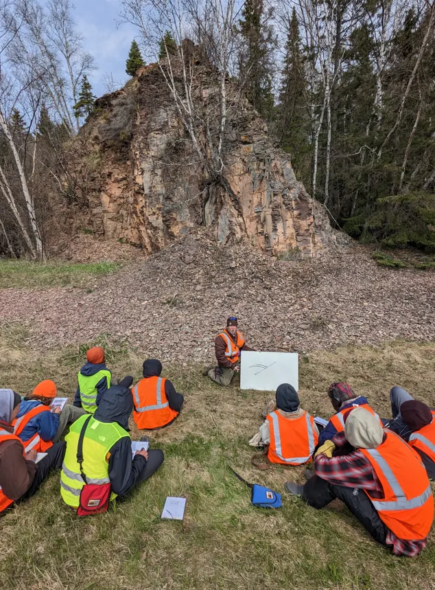 Group of students in reflector vests sit around facing instructor with large rock formation in background.
