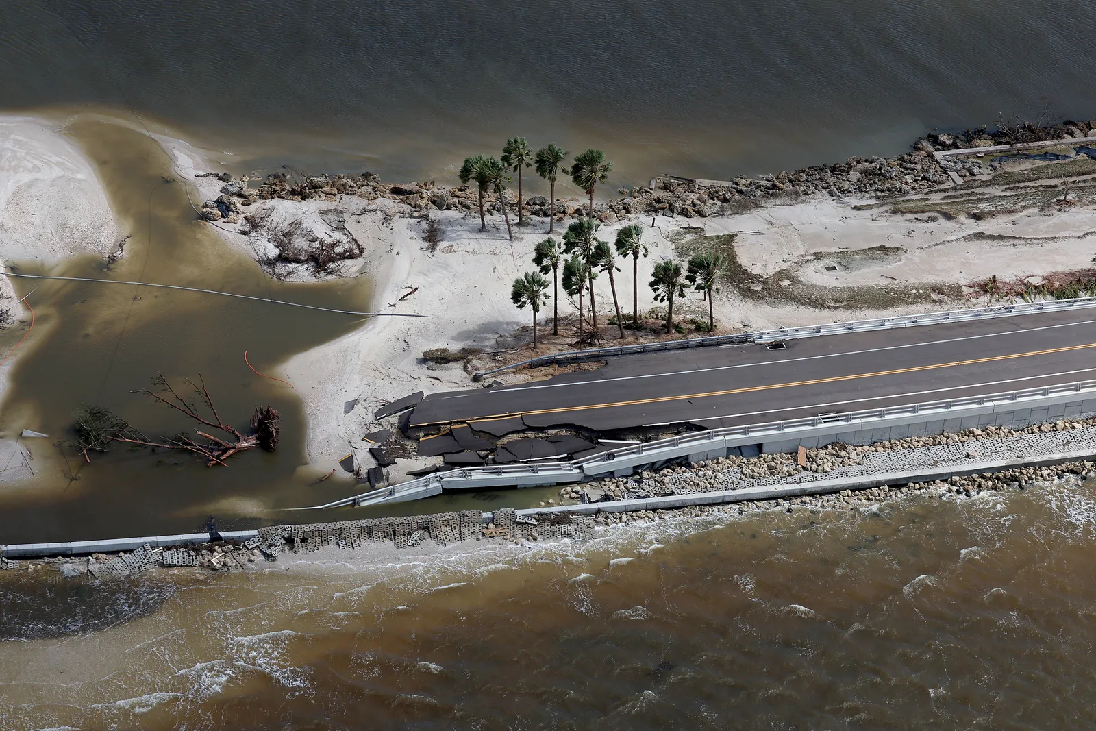 An aerial view shows ocean overtaking a road, which has cracked asphalt and crumpled railings.