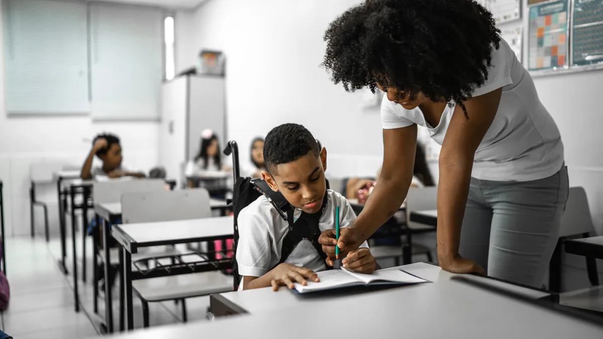 A student wearing a brace on their chest is seated at a desk in a classroom. An adult is standing nearby holding a pencil to a notebook on the desk.