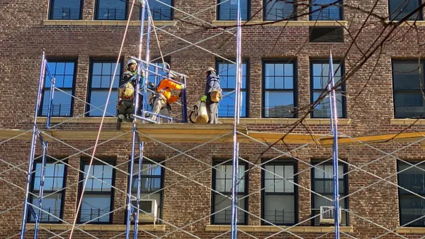 Four people wearing hard hats on scaffolding in front of a brick building.