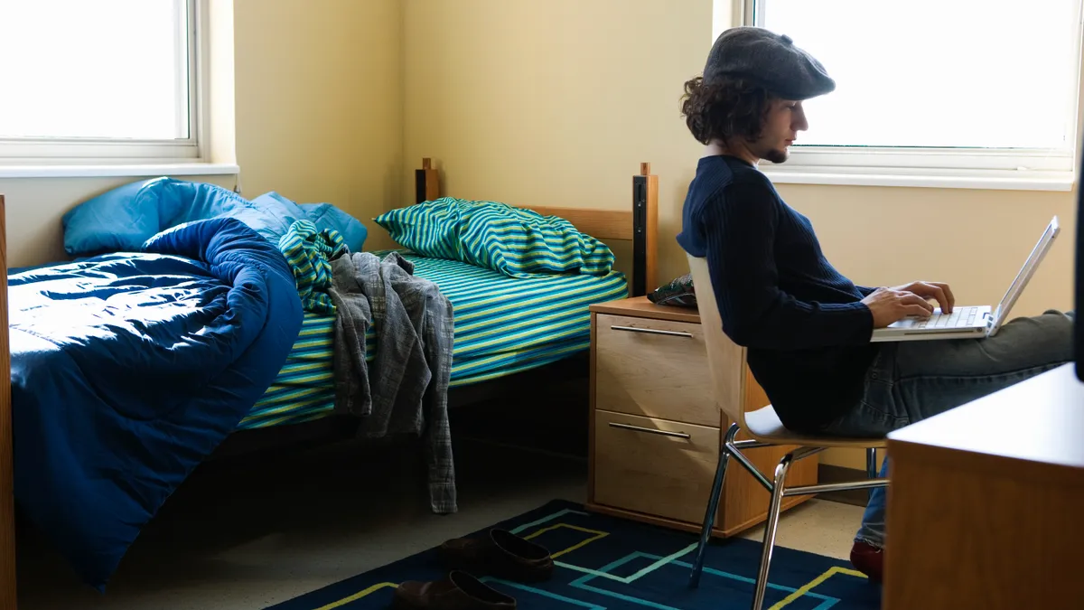 A person sits in a chair with a computer in their lap. An unmade bed is in the background.