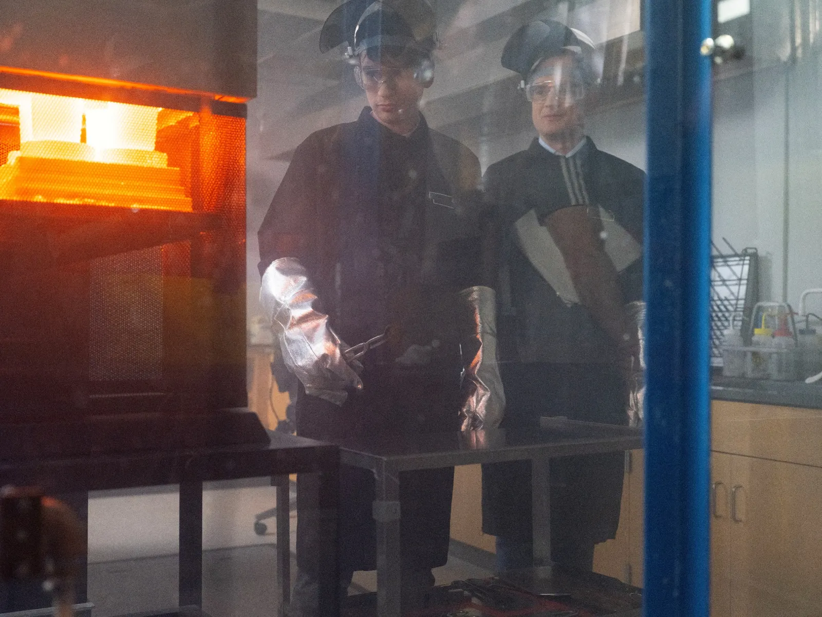 Two people wearing protective gear stand next to equipment containing molten glass.