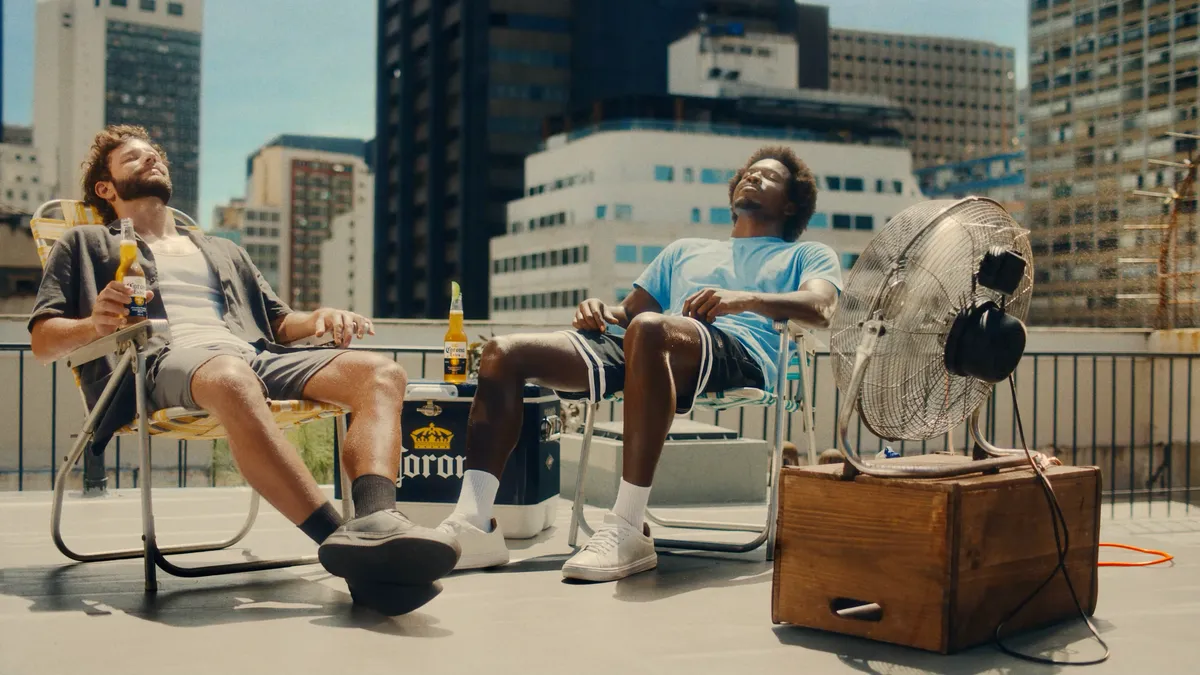 Two men relax in lawn chairs on the roof of a building, one holds a bottle of Corona.