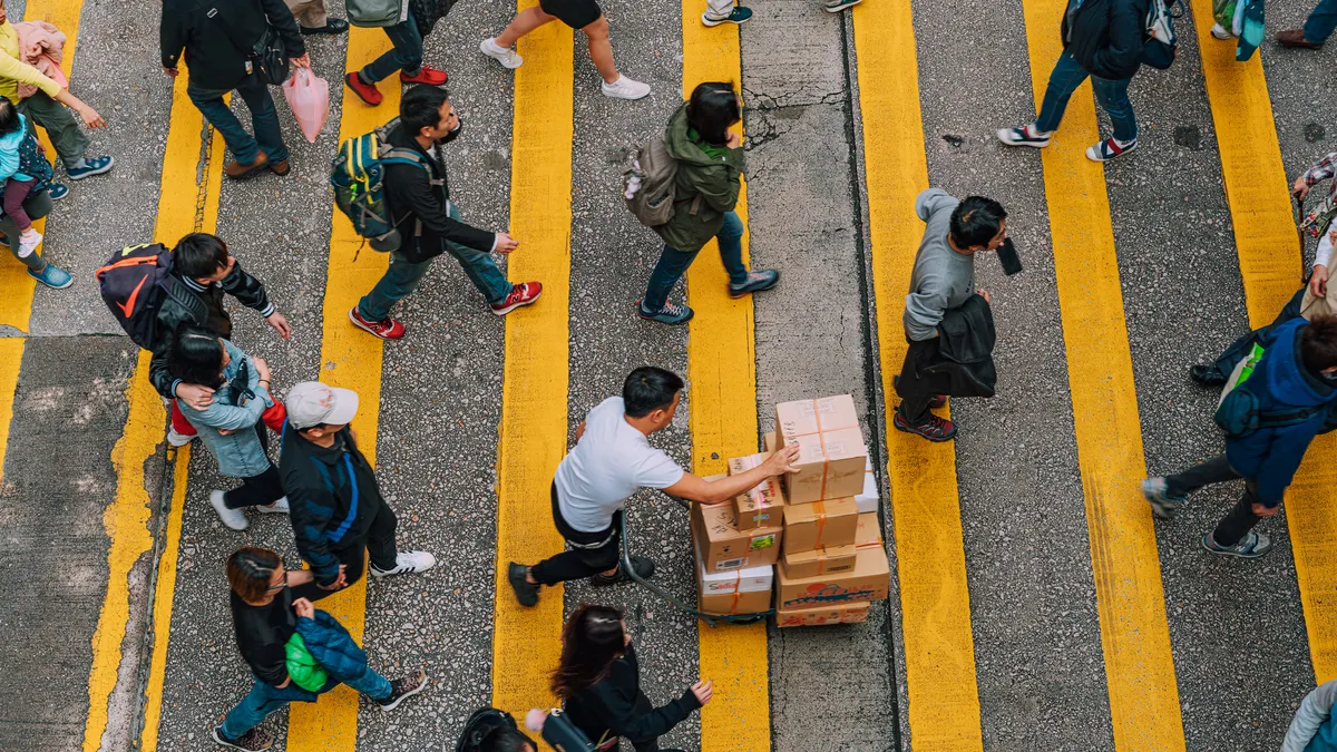 Overhead view of pedestrians and a delivery worker pushing stacked boxes across bright yellow crosswalk lines in busy Hong Kong, capturing urban movement, daily commute and street life energy.