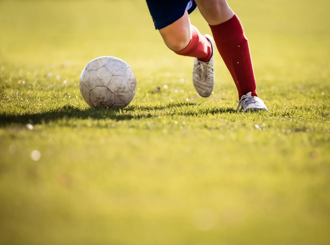 A pair of legs in soccer cleats and red shin guards kicks a soccer ball on a grassy field