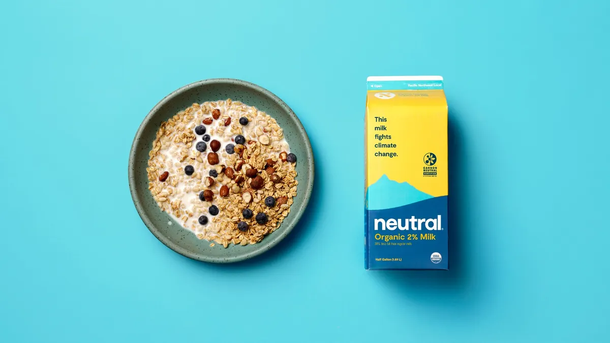A bowl of granola cereal is next to a lying-down carton of Neutral Foods' Organic 2% Milk on a blue background.