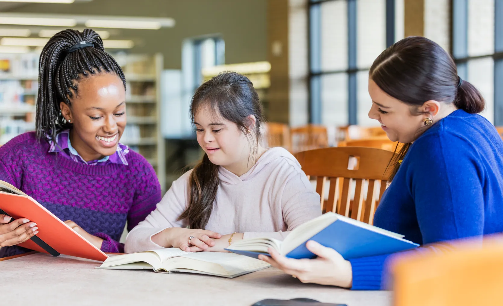 Three older students sit at a desk. They are looking at several open books on the desk.