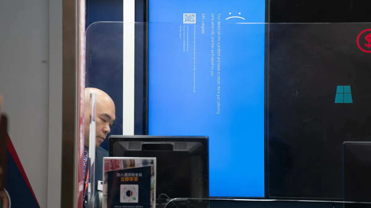 A blue screen with an unhappy face is depicted on a screen in the background behind glass and in front of a person looking down.