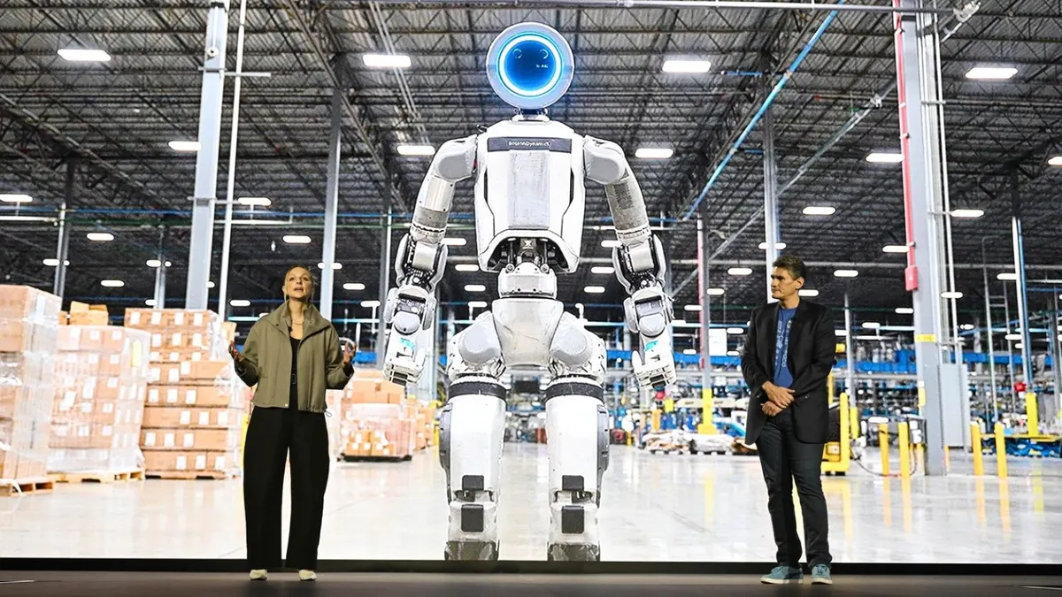 Two people in standing in front of an large high-definition screen of a human robot in a factory.