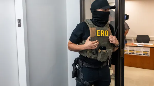 A person wearing a black hat, a black face mask, a vest that says ERO and a gun in a hip holster looks through an open doorway.