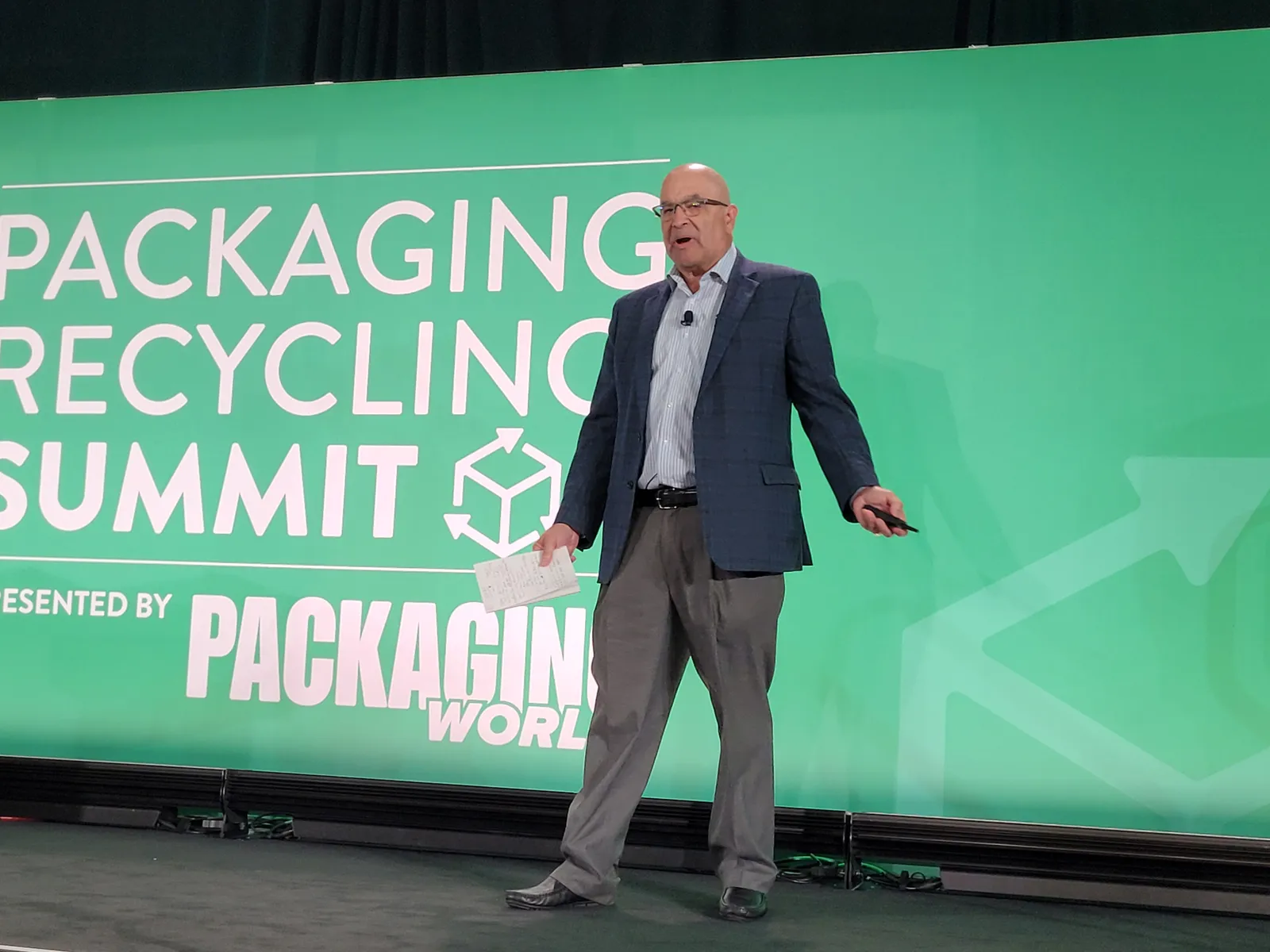 A person on a stage addresses an audience while standing in front of a green background that says Packaging Recycling Summit.