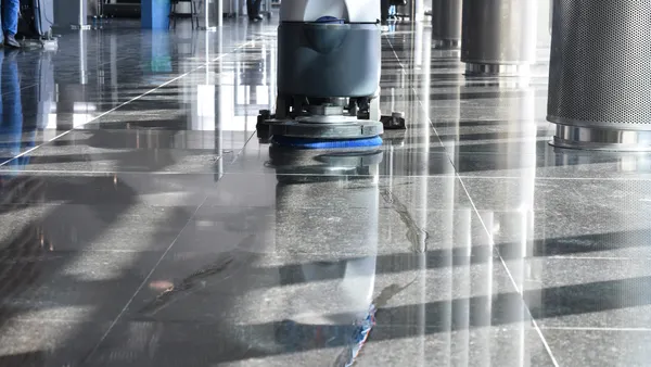 A close-up view of a floor scrubbing machine cleaning a reflective black tile floor in a modern interior with sunlight and shadows.